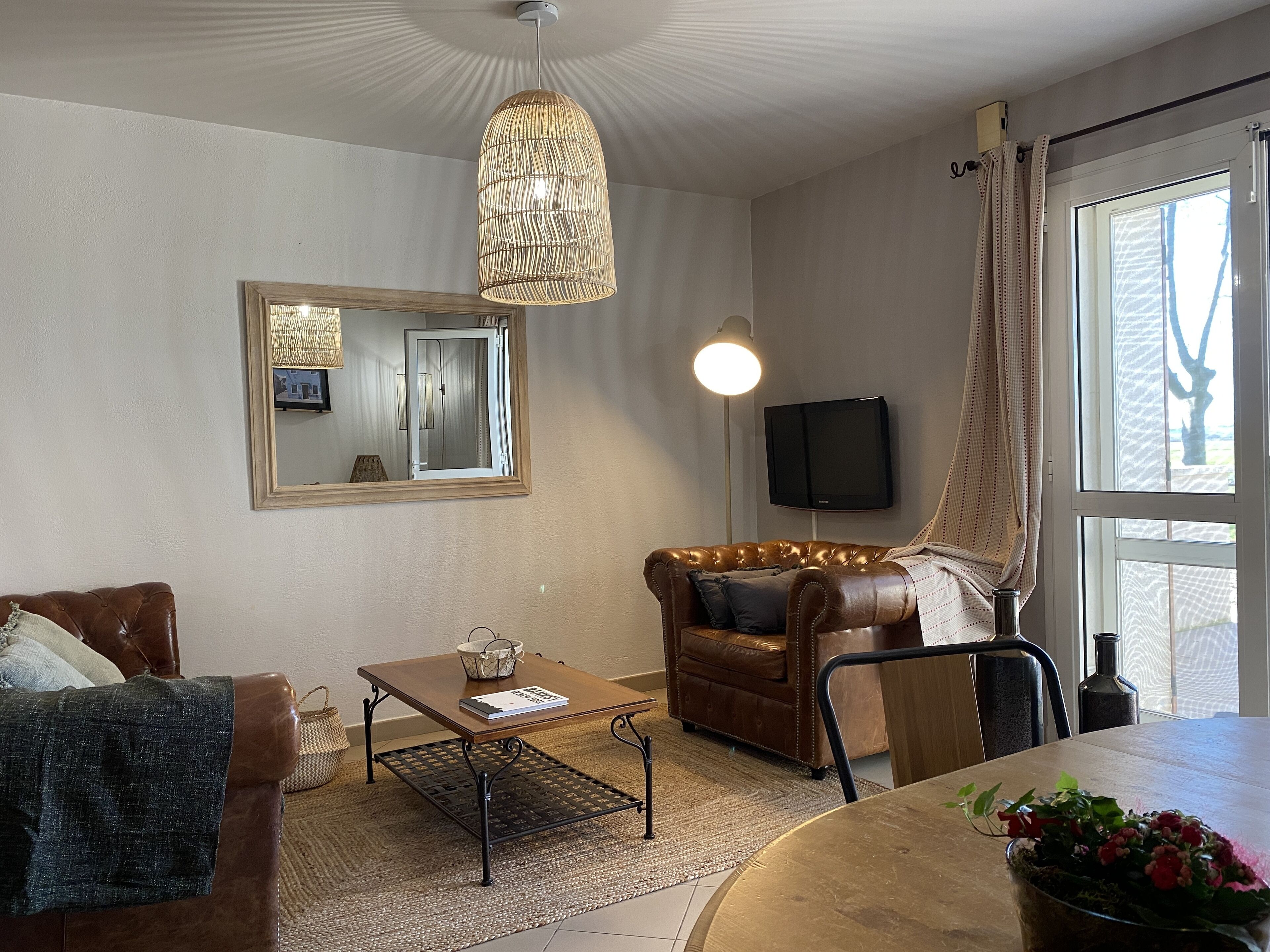 Una sala de estar con un sofá de cuero marrón, una mesa de centro de madera, un espejo, un televisor y una ventana con cortinas.