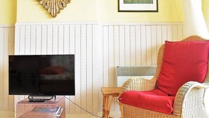 Un salón con televisor de pantalla plana, una silla de mimbre con cojín rojo, un cuadro de paisaje enmarco y un espejo decorativo.