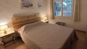 Ett sovrum med en sänggavel i trä, en säng med ett mönstrat överkast, två sängbord med lampor, ett fönster med gardiner och en inramad tavla på väggen.
