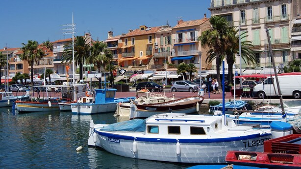 A marina with various boats docked, surrounded by buildings with balconies and palm trees.