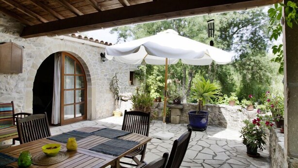 Un patio al aire libre con paredes de piedra, una mesa de madera, sillas y un paraguas grande.