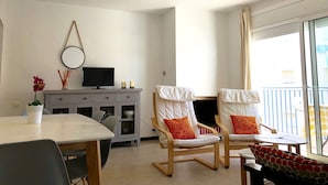 A modern living room with a dining table, two rocking chairs with cushions, a TV on a cabinet, and a large window with a view. - APARTAMENTOS AQUARIUM (REF 31). NEXT TO THE BEACH. RENOVATED. (Roses)