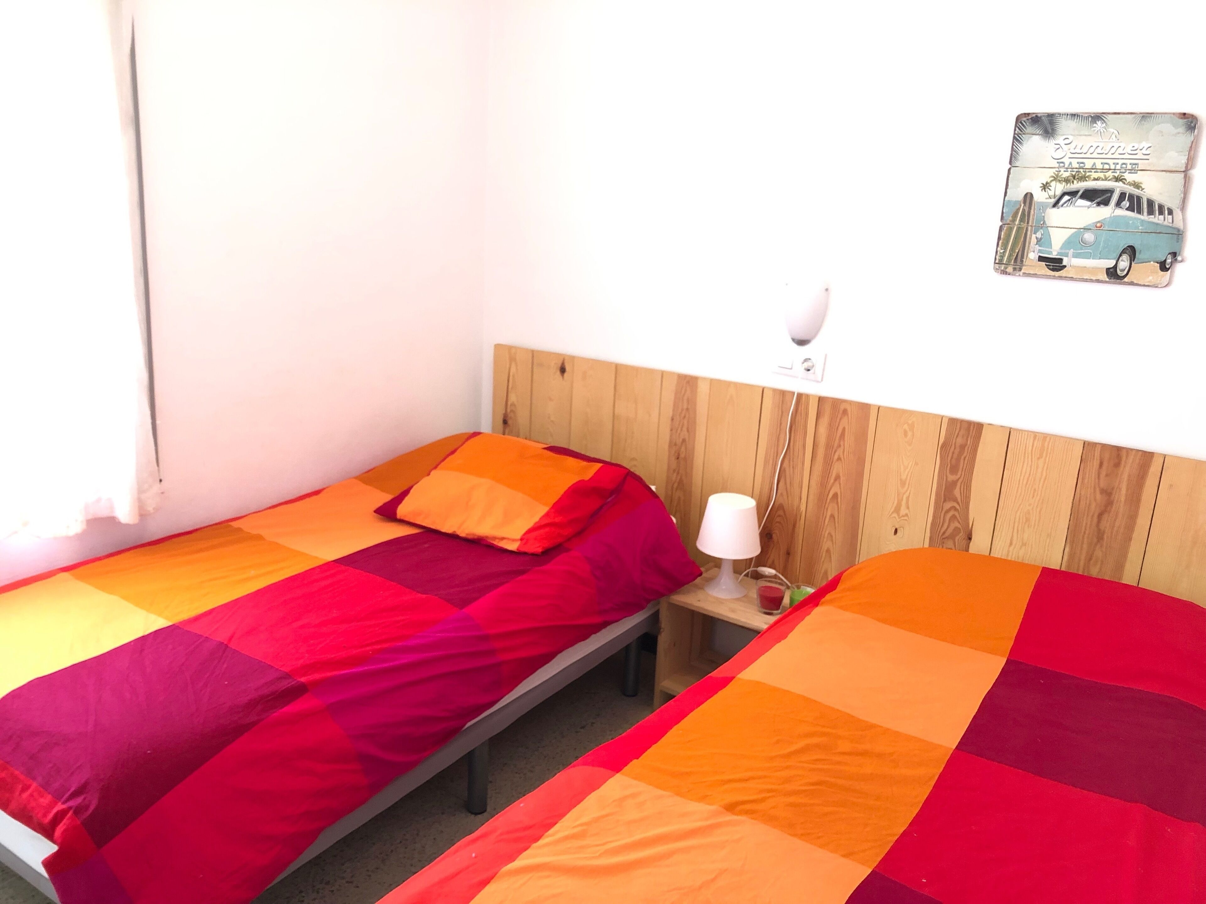 Une petite chambre avec deux lits simples, chacun recouvert d’une couverture colorée. On y trouve une table de chevet avec une lampe et une petite table avec une boisson.
