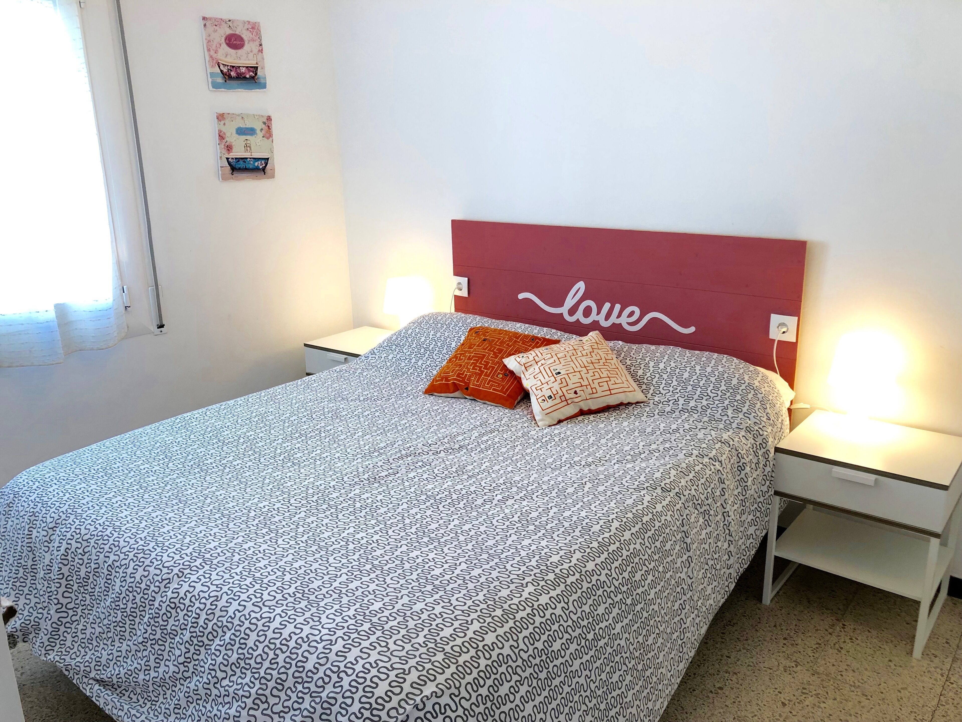 A neatly made bed with patterned bedding, two pillows, a headboard with 'love' written on it, bedside lamps, and a small nightstand.