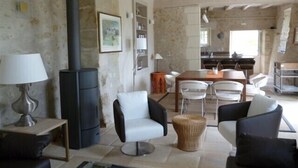 Un interior rústic amb una paret de pedra, una estufa negra, cadires blanques i una taula de dinar.