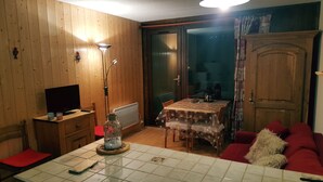Un salon chaleureux avec des murs en bois, une table à manger recouverte d’une nappe à motifs, un canapé rouge et un téléviseur posé sur un meuble en bois.