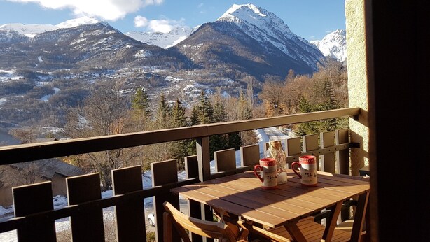 Une terrasse en bois avec une table et des chaises, offrant une vue sur un paysage montagneux aux sommets enneigés.