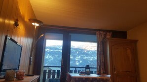Une pièce avec un intérieur en bois, une table sur laquelle se trouvent un gâteau et une tasse, un téléviseur, et une vue sur des montagnes enneigées par une grande fenêtre.