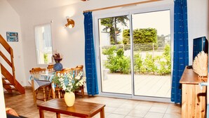 A spacious living area with a dining table, sofa, and a view of the garden through large sliding glass doors. - 40 meters from the BEACH / SEA Quiet house, Private Parking, Charges INCLUDED (Barneville-Carteret)