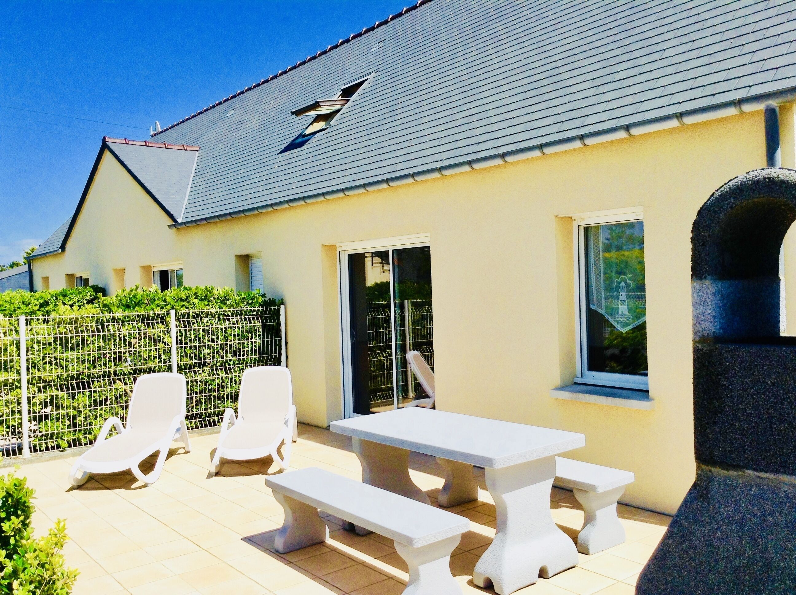 Ein Haus mit einer gefliesten Terrasse, weißen Möbeln und einem umzäunten Garten.