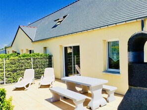 Een huis met een betegeld terras, witte meubels en een omheinde tuin.