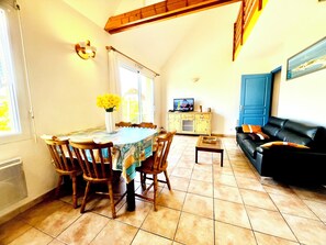 Un coin repas avec une table, des chaises et un vase de fleurs. On trouve un téléviseur posé sur un meuble et un canapé dans le salon.