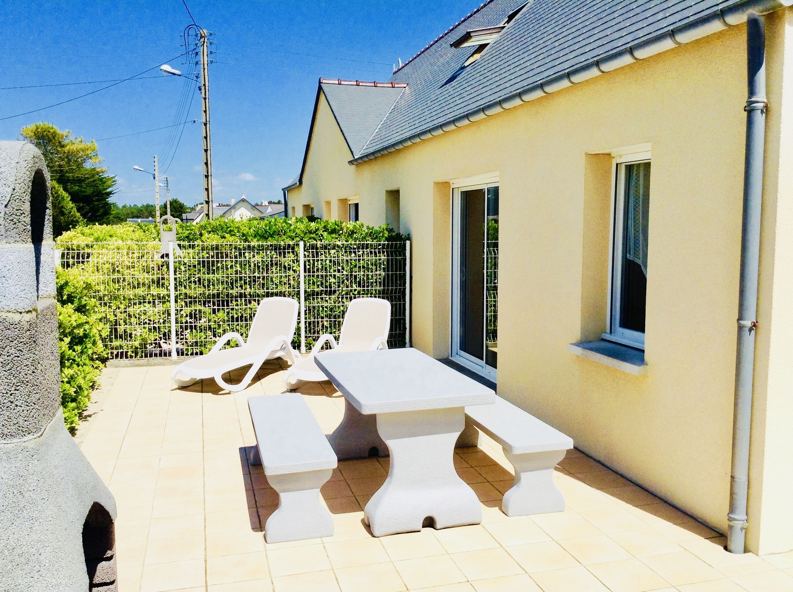 Eine Terrasse mit Tisch und zwei Liegestühlen, ein weißer Zaun und ein Haus mit grauem Dach.