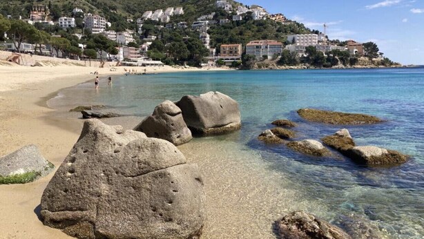 Una playa con agua cristalina, rocas grandes y edificios al fondo.