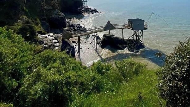 Eine Küstenlandschaft mit einem felsigen Strand, einem hölzernen Steg und einer kleinen, auf Stelzen stehenden Konstruktion.