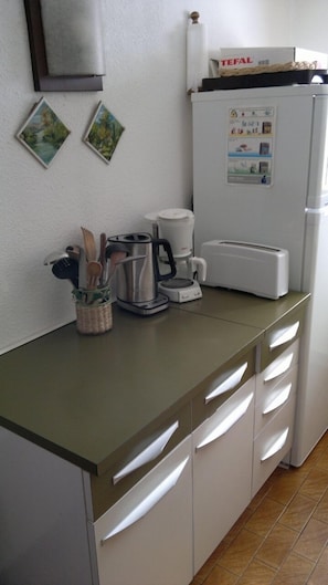 Et køkken med kaffemaskine, brødrister og et køleskab med TEFAL logo. To billeder hænger på væggen, og en kurv indeholder redskaber.