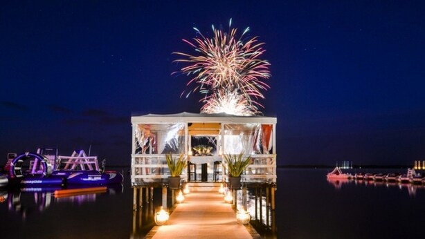 A waterfront venue with a covered seating area, a reflective water path, and boats in the background.