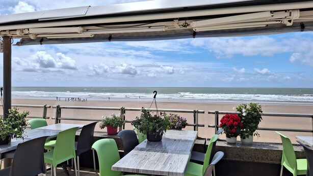 Un espace extérieur aménagé avec des tables et des chaises, offrant une vue sur une plage où l’on aperçoit des vagues et des personnes au loin.