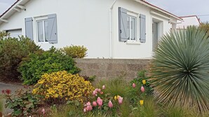 Une maison blanche aux volets gris, entourée d’un jardin où poussent des fleurs jaunes, des roses roses et un grand yucca.