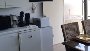 Kompakti keittiö, jossa on valkoiset kaapit, jääkaappi, mikroaaltouuni ja kahvinkeitin. Seinälle, mikroaaltouunin yläpuolelle, on asennettu litteänäyttöinen televisio. Huoneessa on suuri ikkuna, josta on näkymä ulos.