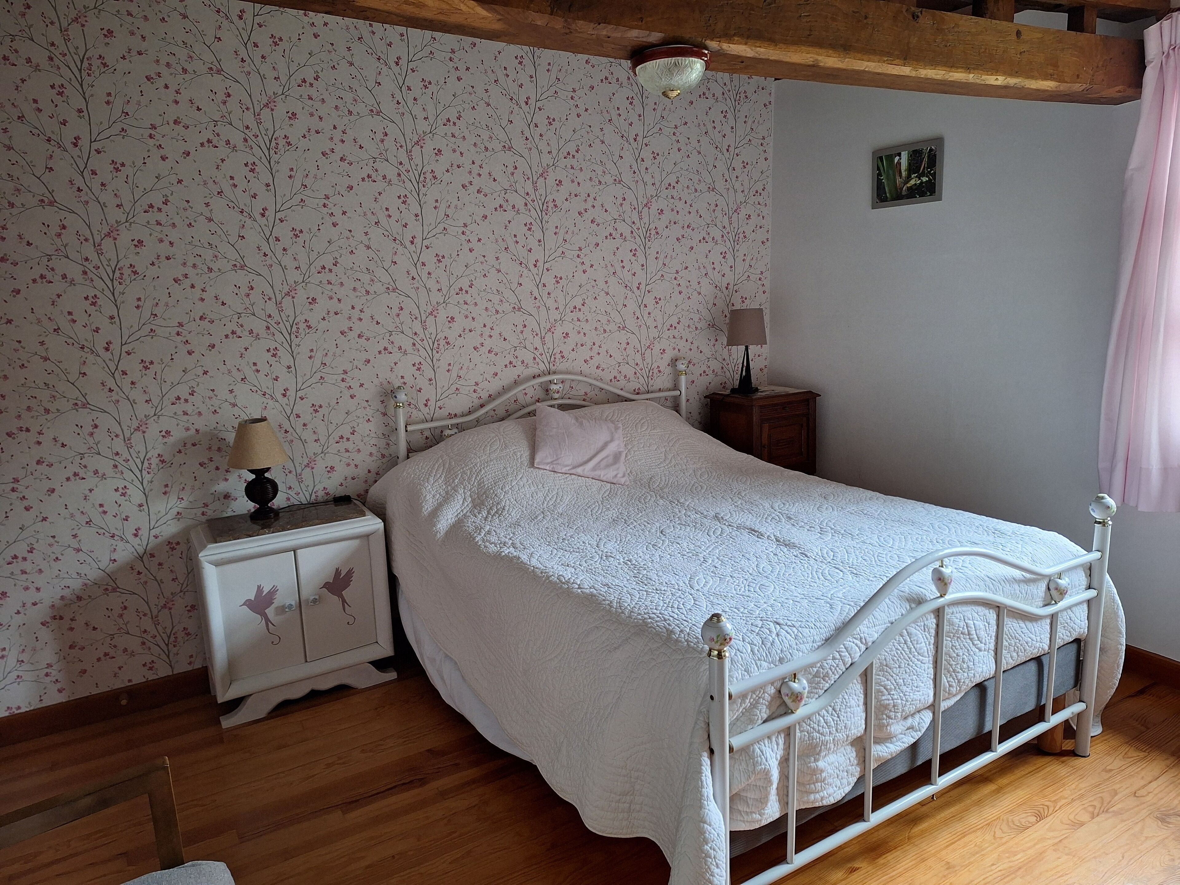 Ein Schlafzimmer mit einem weißen Bett, einem Nachttisch mit Lampe und floraler Tapete.