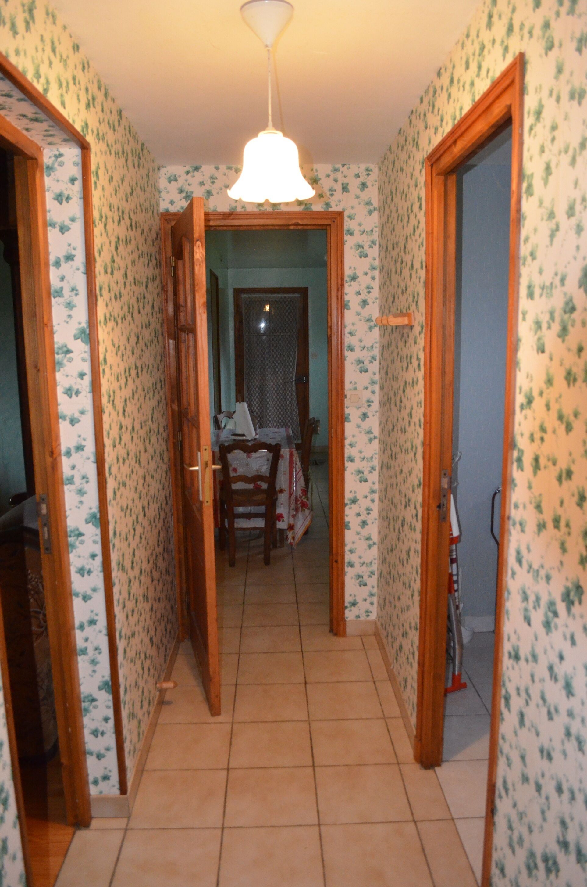 A narrow hallway with patterned wallpaper, wooden doors, and a tiled floor leading to a dining area with a table and chairs.