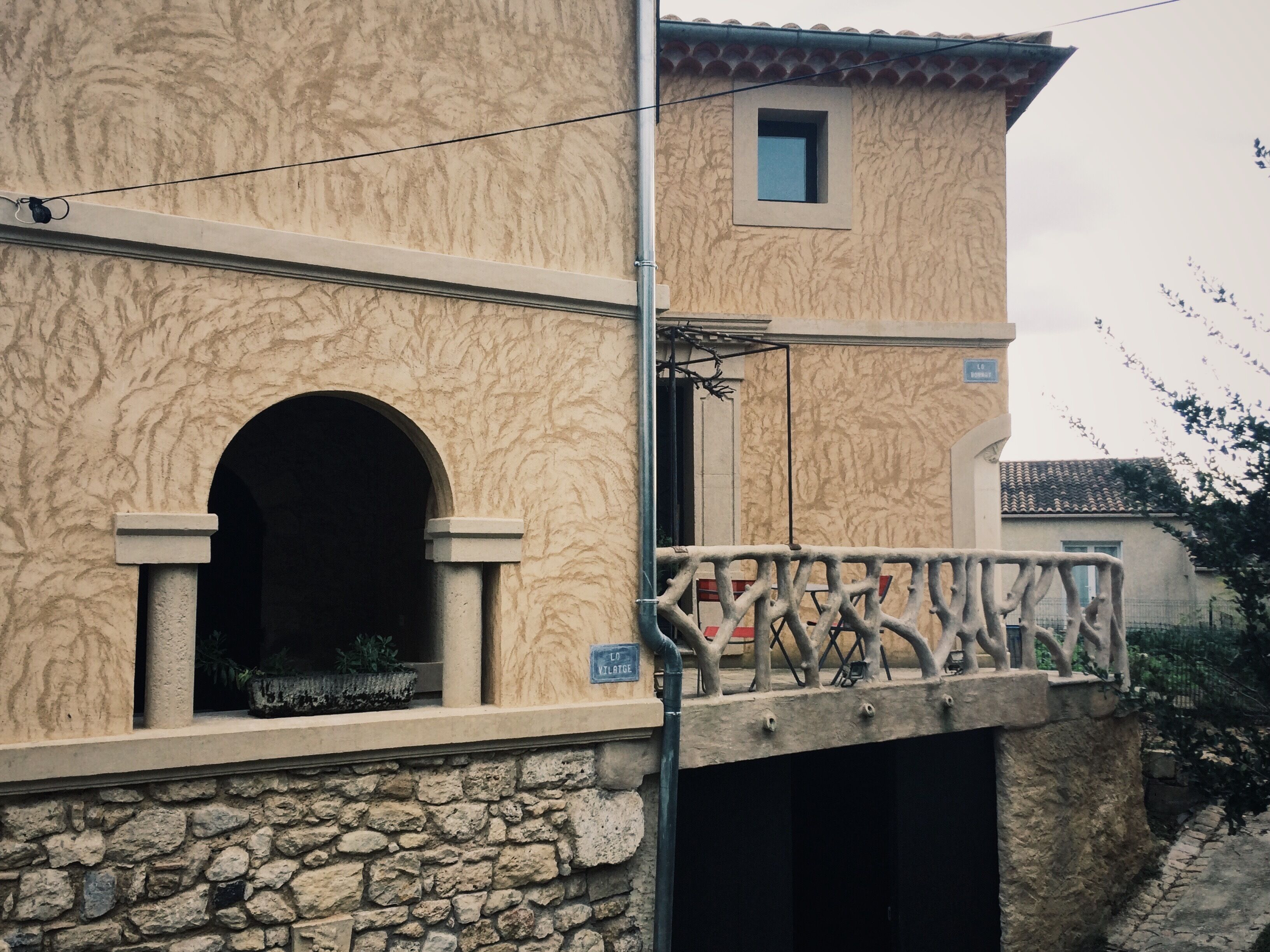 Un bâtiment dont la partie inférieure est en pierre et la partie supérieure de couleur beige, agrémenté d’un balcon doté d’une balustrade en fer forgé.