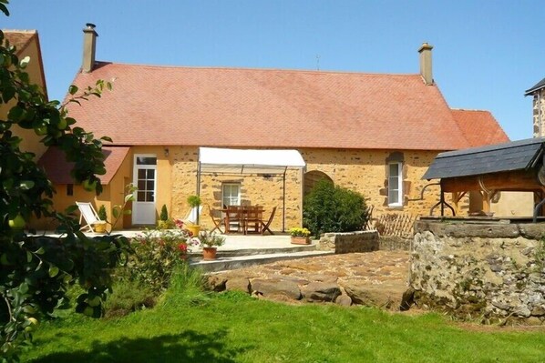 A stone building with a red roof, a covered patio with a table and chairs, and a garden area with a lawn and potted plants.