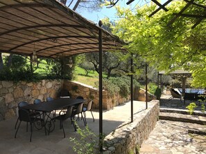 Une terrasse extérieure avec un mur en pierre, une table avec des chaises et une pergola surmontée d’une cloche.