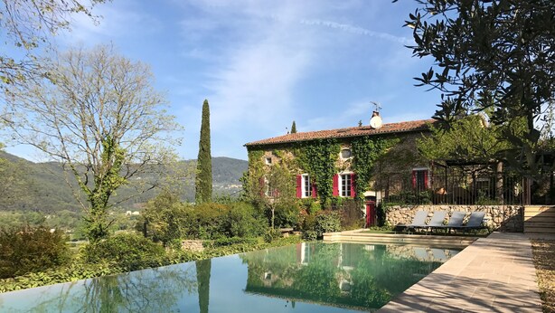 Une piscine entourée d’une terrasse en pierre, d’un mur en pierre et d’un bâtiment aux volets rouges et orné de lierre.