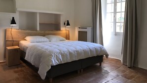 Une chambre avec une tête de lit en bois, un lit avec une literie blanche, deux tables de chevet avec des lampes, une fenêtre avec des rideaux et un radiateur.