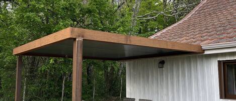 A covered outdoor seating area with a table and chairs, a wooden pergola, and a white building with a tiled roof.