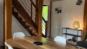 Ruokailutila, jossa on puinen pöytä ja valkoiset tuolit, portaikko ja seinään kiinnitetty valaisin.