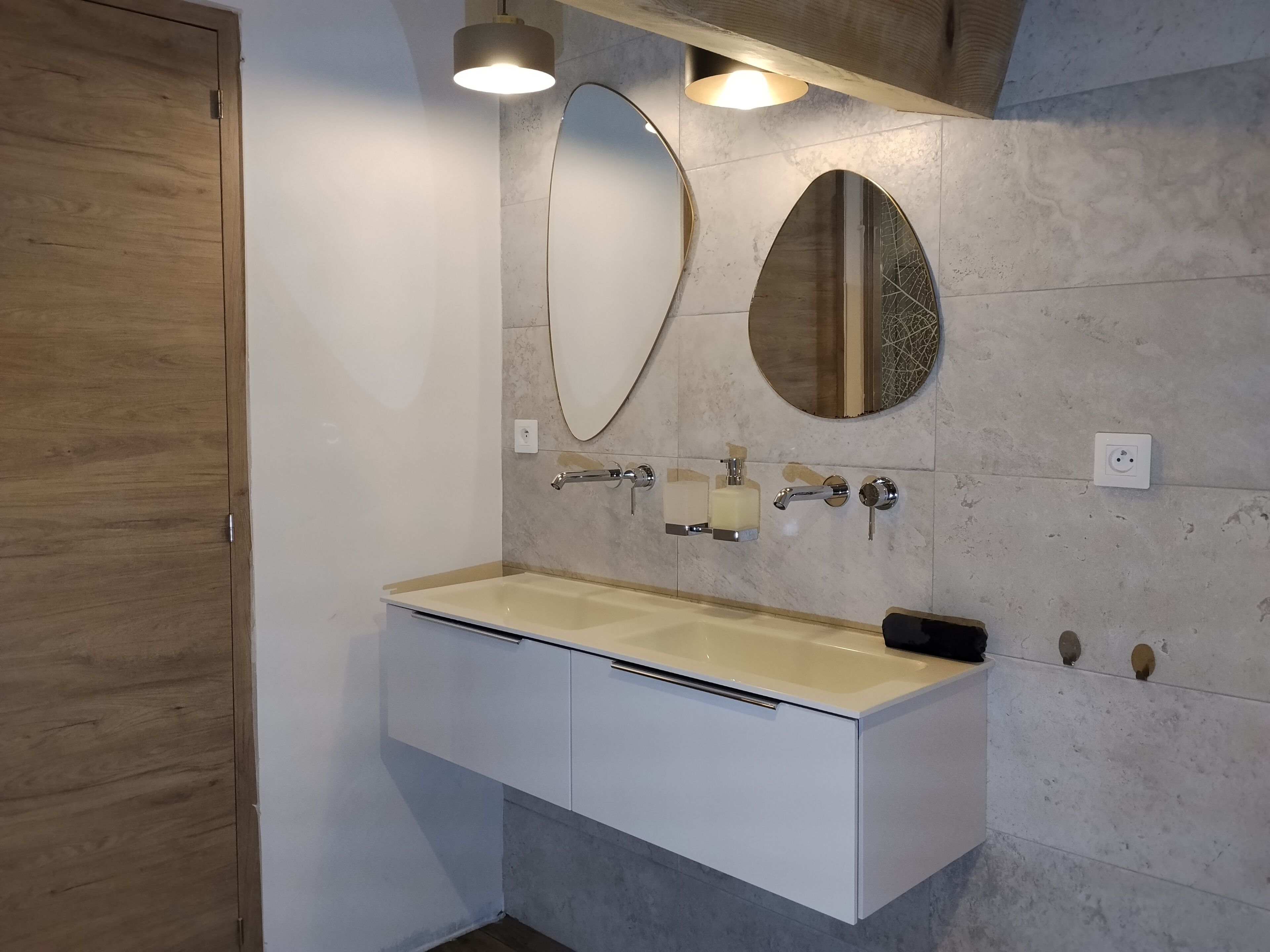 Une salle de bain moderne avec une porte en bois, un meuble-lavabo blanc, un évier beige et deux miroirs fixés au mur.