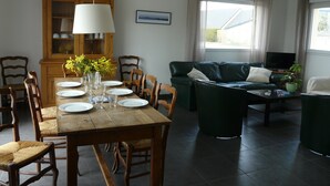 Een eetgedeelte met een houten tafel gedekt voor zes personen, omringd door houten stoelen. Links staat een kast met glazen deuren, en op de achtergrond staat een groene bank met een televisie op een dressoir.