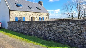 A stone house with a sloped roof and blue shutters, surrounded by a stone wall and a grassy area. - BRETONNE LONGERE BETWEEN SEA AND COUNTRYSIDE (Plouhinec)
