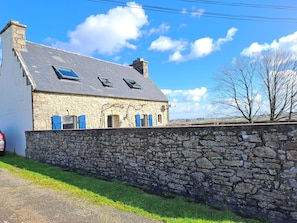 Une maison en pierre avec un toit en pente et des volets bleus, entourée d’un mur de pierre et d’une zone herbeuse.