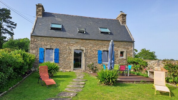 A stone house with a sloped roof, a front door, and a small porch with a chair. - BRETONNE LONGERE BETWEEN SEA AND COUNTRYSIDE (Plouhinec)