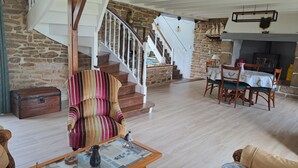 A living room with a striped armchair, a wooden table with a vase, a staircase with stone walls, and a dining area with wooden chairs. - BRETONNE LONGERE BETWEEN SEA AND COUNTRYSIDE (Plouhinec)