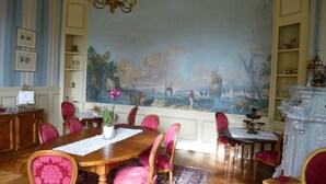 A dining room with a large mural, wooden table, and red upholstered chairs. - Château de la Voûte, facing south (Troo)