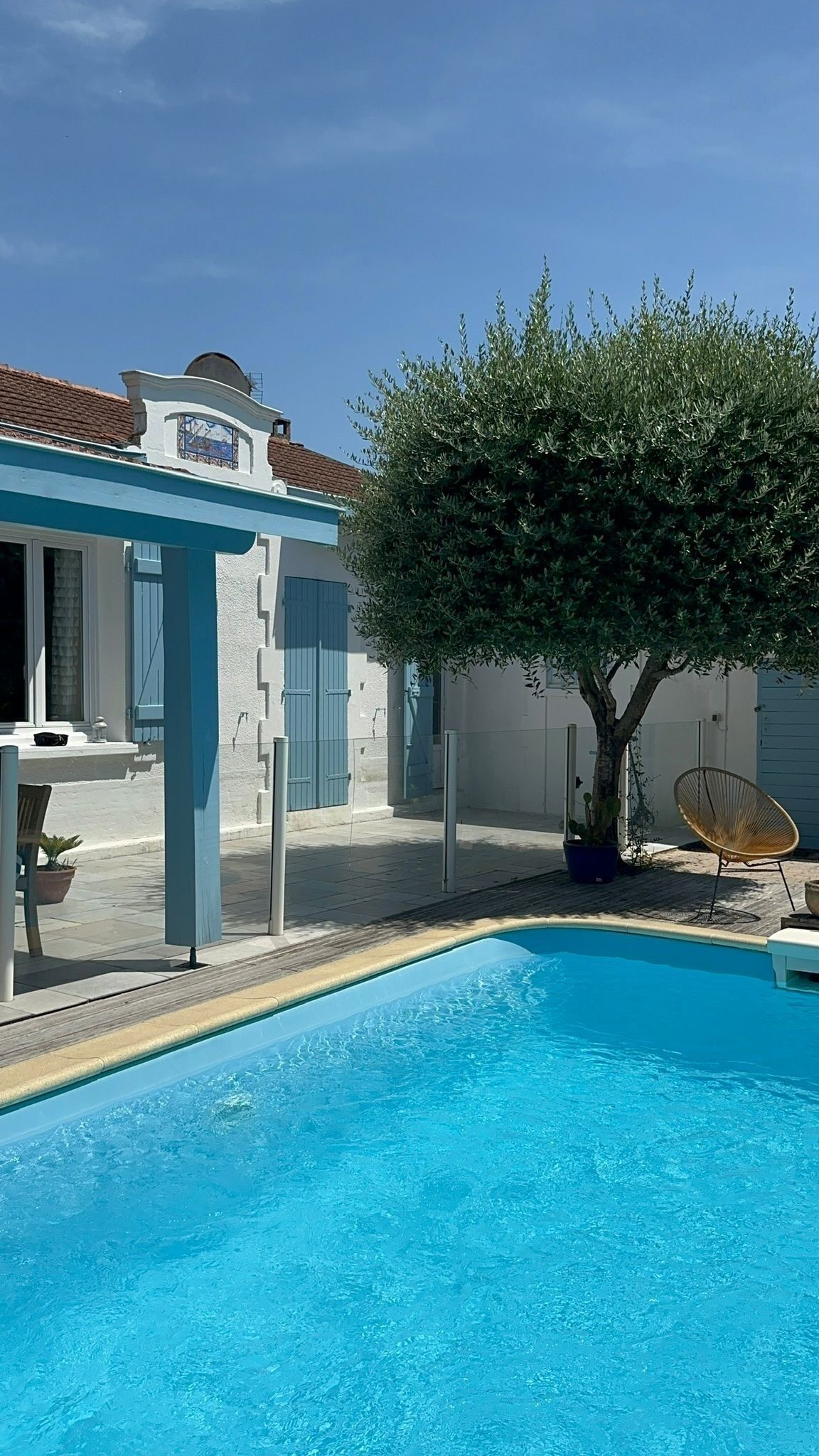 Una piscina con agua azul cristalina, un edificio blanco con contraventanas azules y un árbol al fondo.