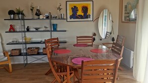 Een eethoek met een houten tafel en stoelen, een klein plankje met decoratieve objecten, en een kunstwerk in een lijst aan de muur.