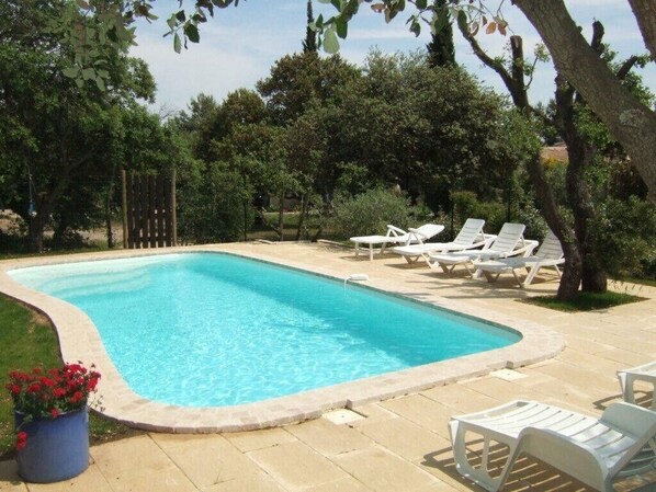 A rectangular swimming pool with a tiled edge, surrounded by a patio area with white lounge chairs and a potted plant with red flowers.