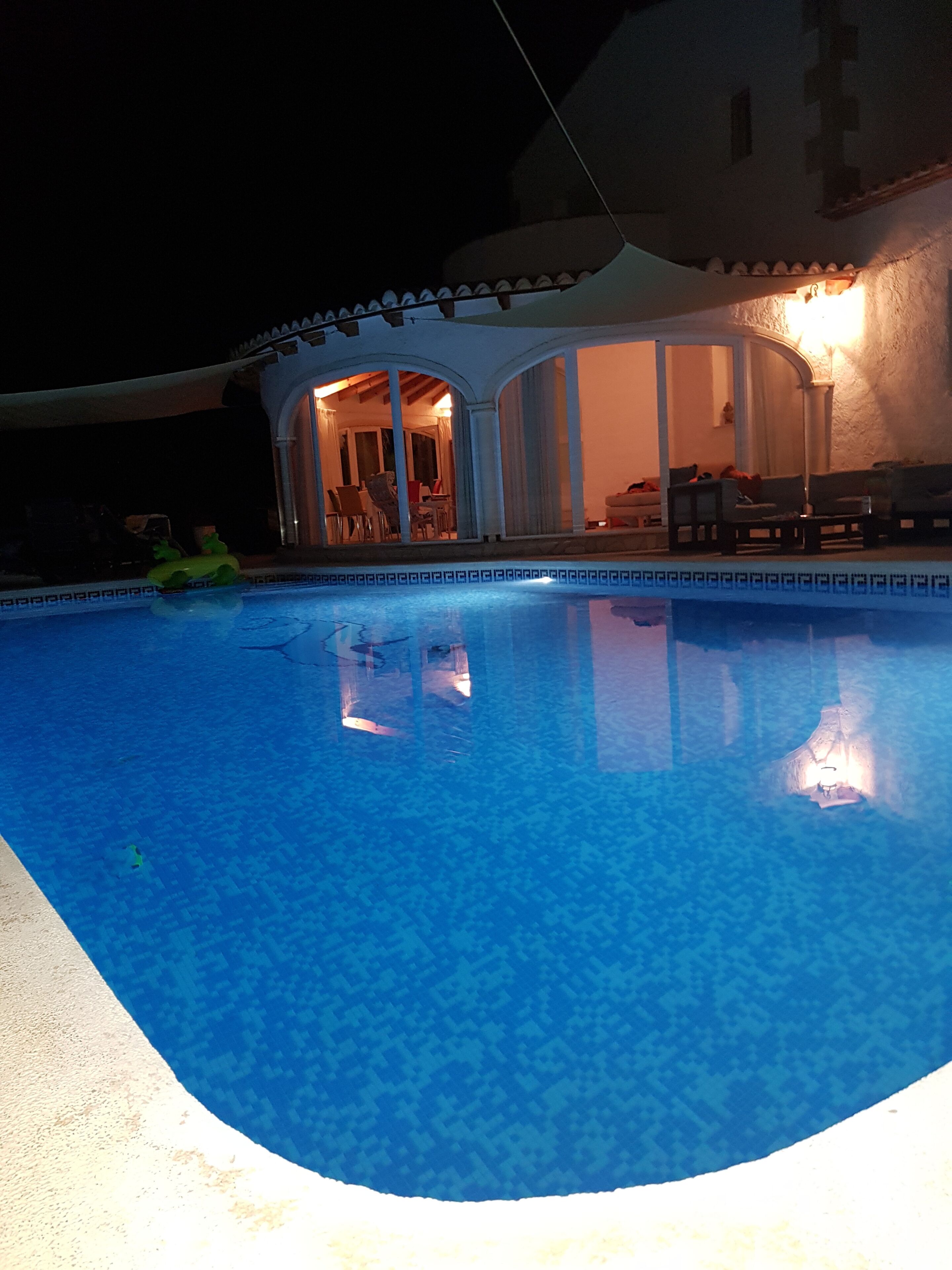 Ein Swimmingpool mit glatter, blauer Wasseroberfläche, umgeben von einer überdachten Terrasse mit Stühlen und einem Tisch.