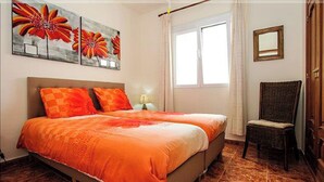 Een tweepersoonsbed met een oranje sprei, een stoel en een raam met gordijnen.