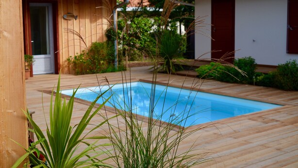 A wooden deck with a swimming pool, surrounded by greenery and a building with a tiled roof. - Superb seaside villa, warm and fully equipped (Vieux-Boucau-Les-Bains)