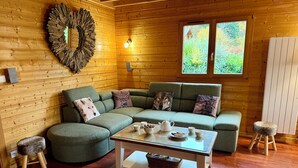 En mysig stuga med en grön, modulbaserad soffa, ett trägolv, en dekorativ spegel som hänger på väggen och ett fönster med utsikt över grönskan.
