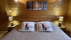 En stuga med träpanel på väggarna, ett rum med en säng som har kuddar med älgtema, en målning av en skog och två sänglampor.