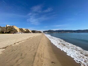 A beach with a sandy shore, a clear blue sky, and buildings in the background. - APARTAMENTOS AQUARIUM (REF 2A). ON THE BEACHFRONT, PERFECT X 2. (Roses)