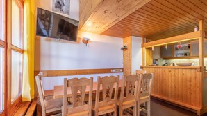 Un coin repas en bois avec une table et des chaises, un téléviseur fixé au mur et un espace cuisine avec des armoires et des étagères en bois.